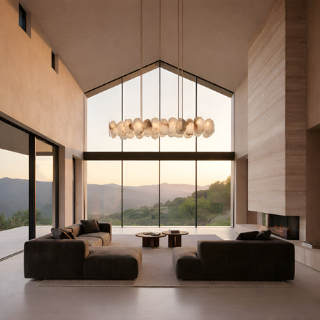 A modern living room with high ceilings, floor-to-ceiling windows framing mountain views, dark sofas, minimalist decor, and the Solvane LED Small Petal Alabaster Linear Suspension Light in soft white & brass hanging overhead.