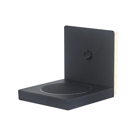 Zanzibar LED wall light in sand black, square matt metal faceplate with backlit edge glow, circular push switch, and integrated wireless phone charging shelf below, shown on a plain white background.