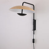 Niori Ginger LED Wall Light in black and brown, shown on a plain wall with a curved metal arm, wood-tone disc shade, black under-bowl, and inline cord switch.