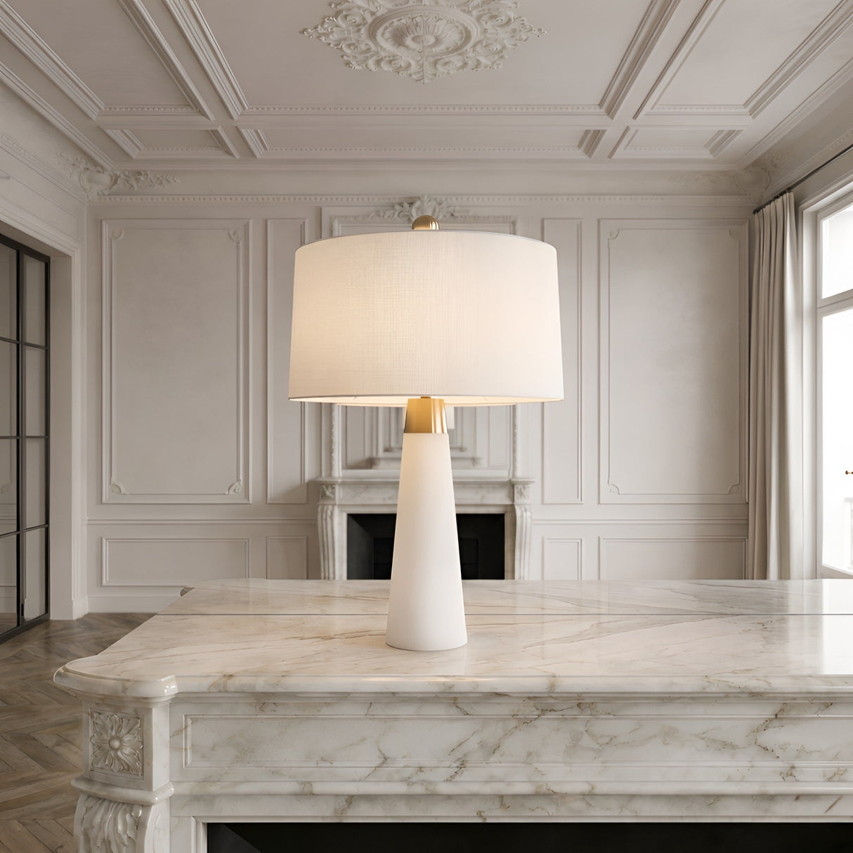 The Arvello 1 Light Slim Tapered Marble Table Lamp in brass and white sits on a marble countertop in an elegant, bright room with ornate wall moldings and large windows.