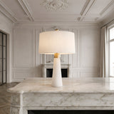 The Arvello 1 Light Slim Tapered Marble Table Lamp in brass and white sits on a marble countertop in an elegant, bright room with ornate wall moldings and large windows.