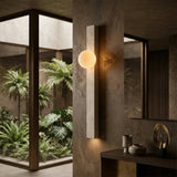 The Selvara 1 Light Vertical Bar Travertine Wall Light in light beige glows on a textured wall near a mirror and decor, with large windows showcasing vibrant indoor greenery.