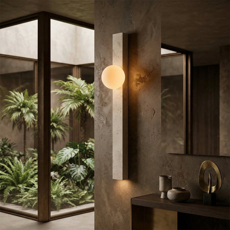 The Selvara 1 Light Vertical Bar Travertine Wall Light in light beige glows on a textured wall near a mirror and decor, with large windows showcasing vibrant indoor greenery.