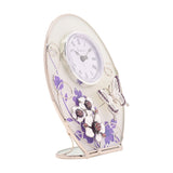 Oval glass mantel clock with Roman numeral dial, purple floral print, silver scrollwork, and a raised butterfly with crystal-centred white flowers, on a plain white background.