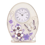 Oval glass mantel clock with Roman numeral dial, purple floral print, raised white butterfly and crystal-centred flowers, silver scrollwork, on a plain white background.