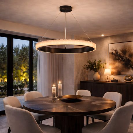 A modern dining room features a round wooden table, four upholstered chairs, and the Lucenza LED Small Halo Ring Alabaster Chandelier in Matte Black & Soft White overhead. Candles, décor accents, and large windows reveal a glowing dusk garden.