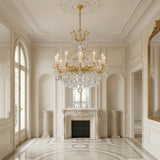 Elegant, sunlit room with cream walls, ornate molding, marble fireplace, and arched windows; a Vienna 12 Light K9 Crystal Chandelier in heirloom gold sparkles above as a gilt mirror reflects the classic French-style decor.
