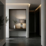 A modern minimalist interior features a built-in wooden shelf displaying the Torvani 1 Light Statement Marble Table Lamp in brass and grey, alongside a ceramic vase with green branches, against gray walls and dark tiled flooring.
