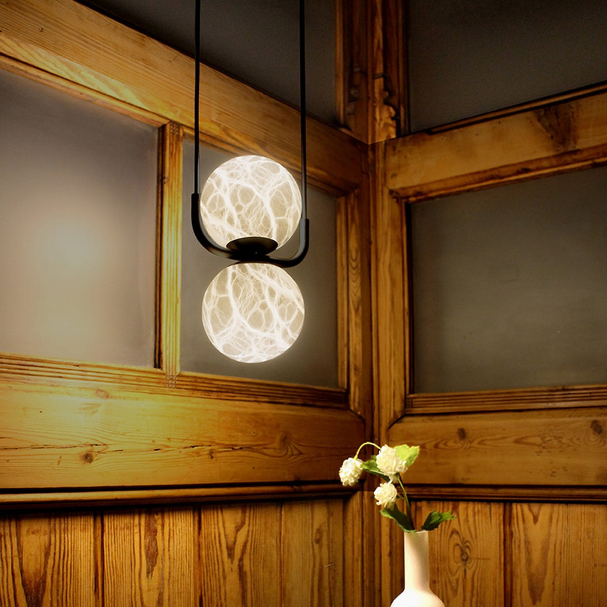 Halvessa LED 2 Light Alabaster Pendant Light in black, two stacked glowing globes on a slim U-shaped iron cradle, shown suspended in a wood-panelled room.