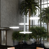 SuperMoon LED 4 Light Pendant Light in white, with a central stem and staggered round crescent-cut discs, shown illuminated above indoor plants in a lobby setting.