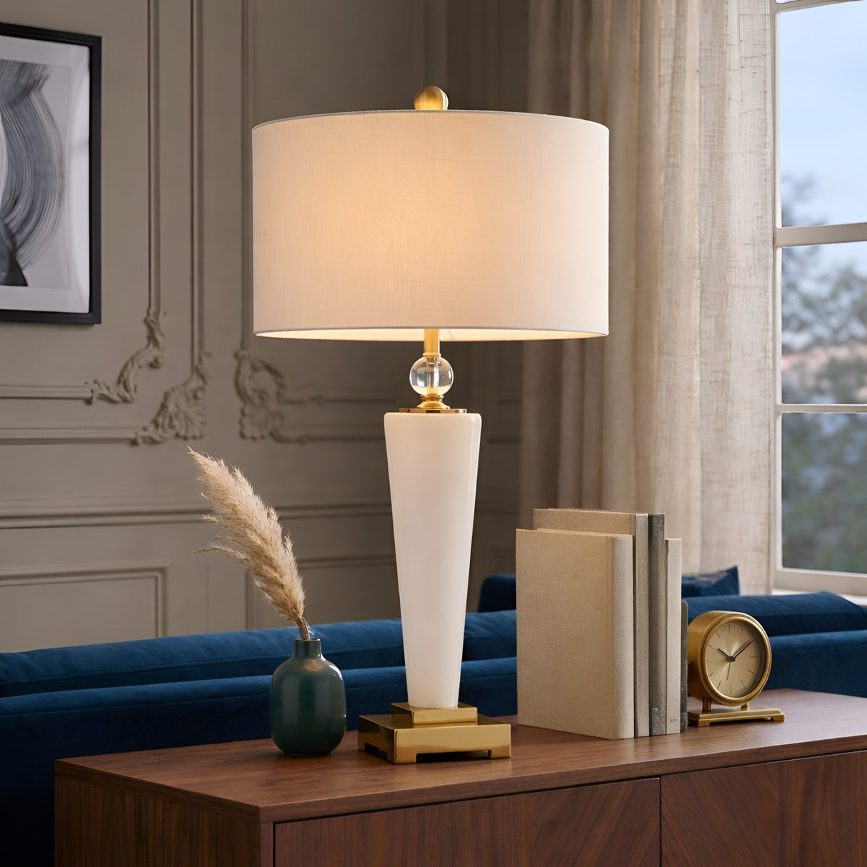 A modern living room features the Aureum 1 Light Refined Silhouette Marble Table Lamp in brass and white on a wooden console, beside stacked books, a green vase with pampas grass, and a gold clock. Elegant molding and sheer-curtained window complete the scene.