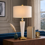 A modern living room features the Aureum 1 Light Refined Silhouette Marble Table Lamp in brass and white on a wooden console, beside stacked books, a green vase with pampas grass, and a gold clock. Elegant molding and sheer-curtained window complete the scene.