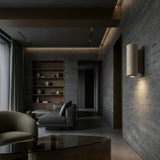 A modern living room with gray textured walls, walnut brown built-in shelves, a gray sectional, floor-to-ceiling curtains, and the Dorsa 1 Light Small Cylinder Travertine Wall Light in Walnut Brown & Light Beige for a minimalist touch.