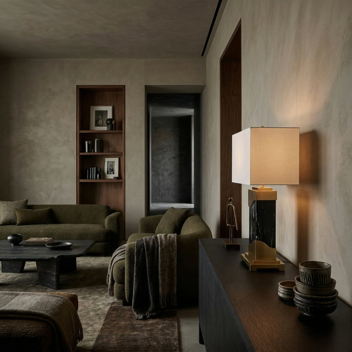 A modern living room features olive green sofas, a dark coffee table, built-in shelves with books, a cozy throw blanket, and a sideboard topped with the Torvani 1 Light Statement Marble Table Lamp in Brass & Black and stacked decorative bowls.