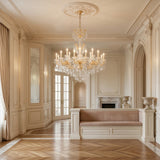 An elegant, cream-colored room features ornate wall paneling, the Vienna 24 Light K9 Crystal Chandelier, large draped windows, a fireplace, and a cushioned built-in bench on a polished herringbone wood floor.