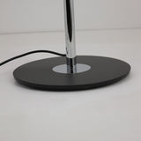 A close-up shows the base of the Stealth 6 Light Floor Lamp in Brass & Black, featuring a black circular platform, chrome pole, and black electrical cord extending from the base on a white surface.