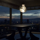 A modern dining area features the Lava Large LED Pendant Light in silver above the table, with two chairs facing each other and large windows showcasing a dusky city skyline in deep blue and orange hues for an inviting atmosphere.