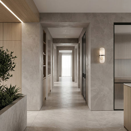 A modern hallway with light gray stone surfaces includes built-in shelves, a planter with greenery, and the Ciel LED Cylinder Alabaster Wall Light in Soft White & Matte Black near frosted glass doors, softly illuminating sheer curtains at the sunlit window.