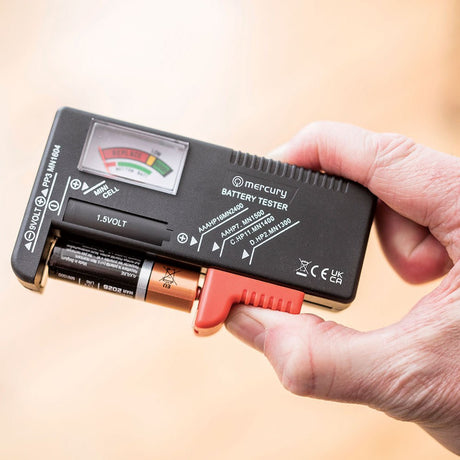 Hand holding the Mercury Universal Analogue Battery Tester by AVSL, a compact black unit with colour-coded needle dial, red slider and AA battery inserted.