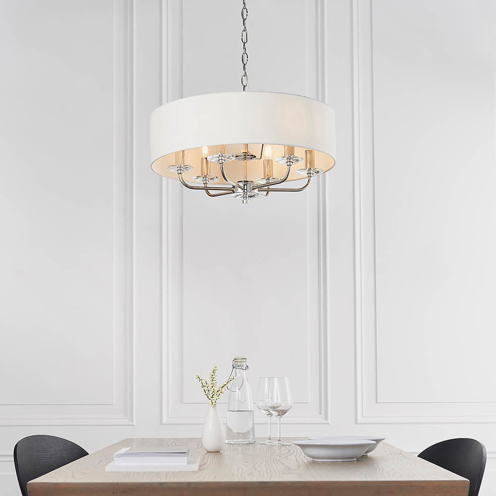 Castdon 6 Light Pendant in nickel with a white drum shade, crystal accents on chrome arms, hung above a dining table in a panelled white room.