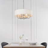 Castdon 6 Light Pendant in nickel with a white drum shade, crystal accents on chrome arms, hung above a dining table in a panelled white room.