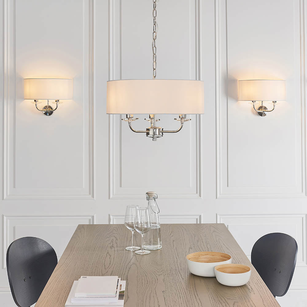 Castdon 2 Light Wall Light in nickel with white semi-circular fabric shade, shown either side of a matching pendant in a dining room setting.