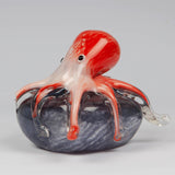 Glass octopus figurine with red and white streaked head and arms draping over a swirled grey rock base, clear glass tentacle tips, on a plain light background.