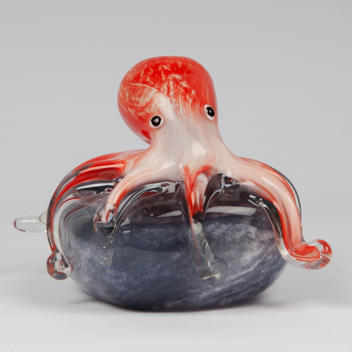 Glass octopus figurine in red and white with clear tentacle tips, draped over a swirled grey rock base, set against a plain light background.