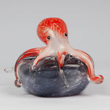 Glass octopus figurine in red and white with clear tentacle tips, draped over a swirled grey rock base, set against a plain light background.