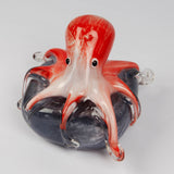 Glass octopus figurine with red and white head, curling tentacles with clear glass tips, draped over a swirled grey rock base, on a plain white background.