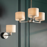 Bexley 3 Light Pendant in matt nickel with curved arms and white fabric drum shades, shown alongside a matching wall light in a dark green panelled room.
