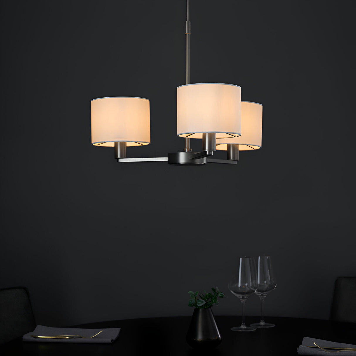 Bexley 3 Light Pendant in matt nickel with three curved arms and white faux silk drum shades, shown lit above a dark dining table.