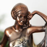 Close-up of the Lady in Draped Floral Gown resin figurine in bronze, showing her face, headband with floral detail, and one hand raised to her temple against a plain light background.