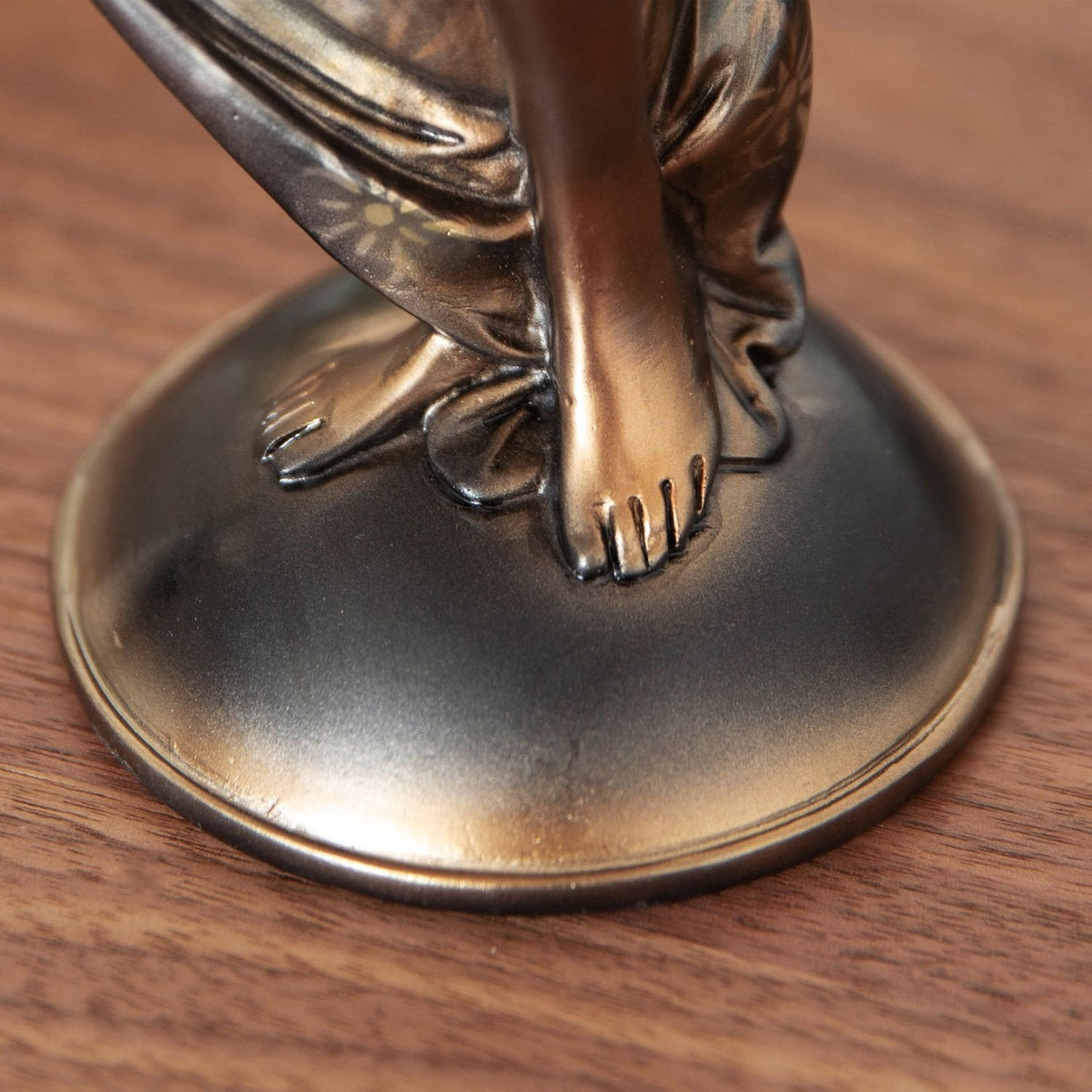 Close-up of the Lady in Draped Floral Gown resin figurine showing bare feet beneath a bronze floral-patterned gown on an oval base, set on a wooden surface.
