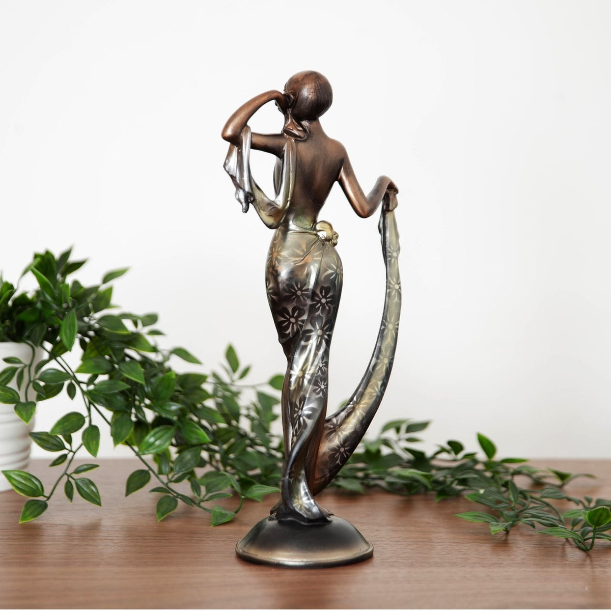 Bronze resin figurine of a woman in a floral draped gown, one hand at her temple and the other lifting her train, shown on wood with greenery.