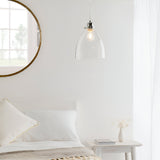 Endon Brackmere clear glass pendant light with polished chrome gimbal and exposed filament bulb, hanging beside a round mirror above a white bed.