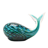 Glass whale figurine with swirled green, turquoise, and navy tones, a raised clear-tipped tail, and a rounded belly, set against a plain white background.