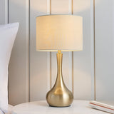 Endon Chartbury LED table lamp with a brass teardrop base and beige drum shade, lit on a bedside table beside a pillow and panelled wall.