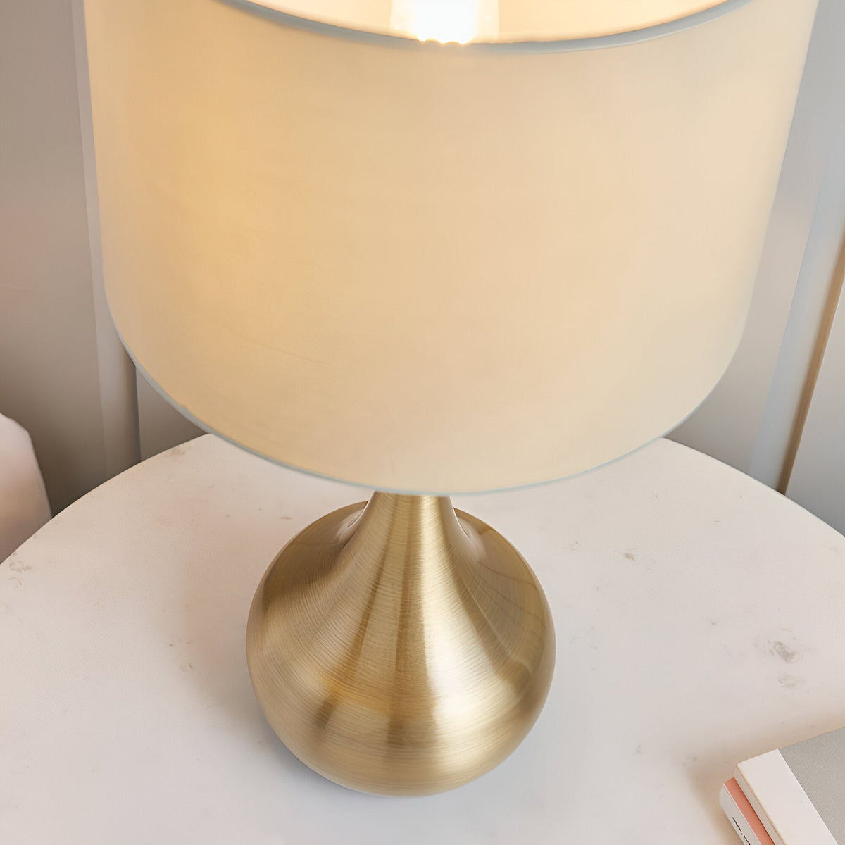Endon Chartbury LED table lamp with brushed brass rounded base and beige drum shade, illuminated on a white bedside table.