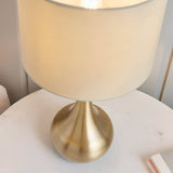 Endon Chartbury LED table lamp with brushed brass rounded base and beige drum shade, illuminated on a white bedside table.