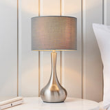 Endon Chartbury LED table lamp with satin nickel curved base and grey fabric drum shade, lit on a marble bedside table beside a pillow.