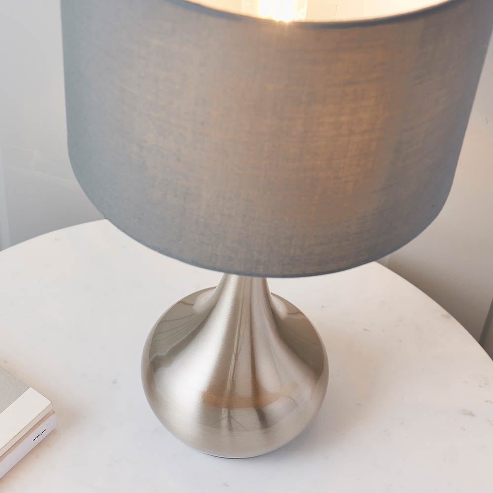 Endon Chartbury LED table lamp close-up showing the satin nickel rounded base and grey fabric drum shade on a white marble side table.