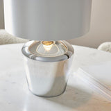 Danefield Table Lamp in chrome, close-up of the rounded glass base with lit E27 bulb beneath a cream fabric shade, set on a marble table.