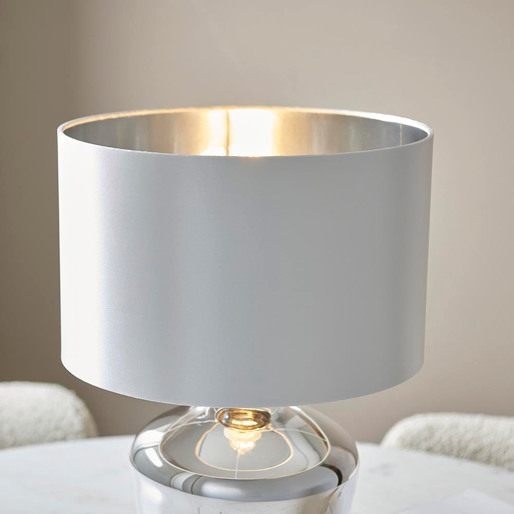 Danefield Table Lamp in chrome and cream, featuring a rounded glass base and cylindrical fabric shade, lit on a marble tabletop.