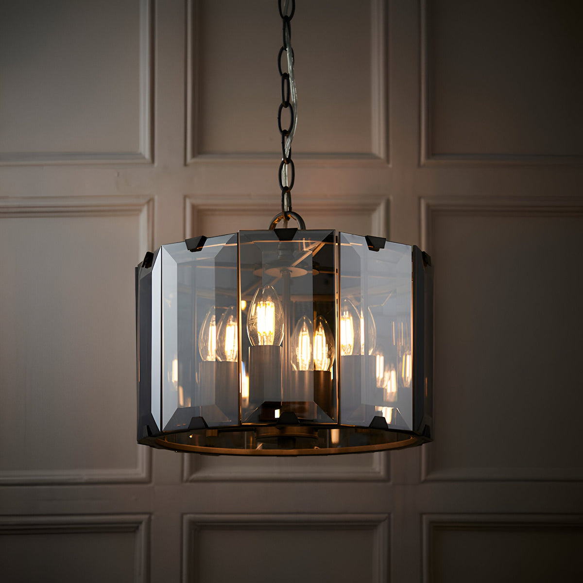 Bevshaw 4 Light Pendant in slate grey, drum shape with bevelled smoke glass panels, hung on a chain, visible bulbs, against a panelled wall.