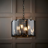 Bevshaw 4 Light Pendant in slate grey, drum shape with bevelled smoke glass panels, hung on a chain, visible bulbs, against a panelled wall.