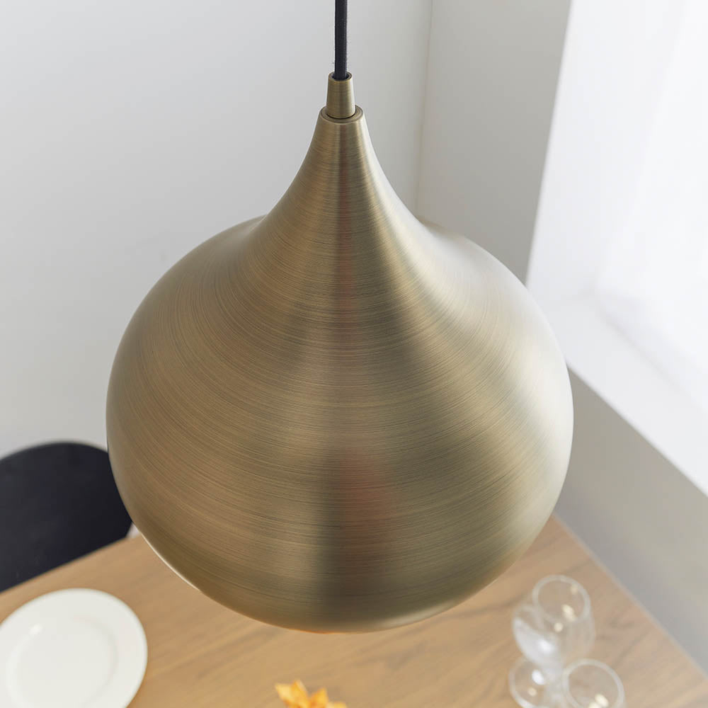Endon Ashleigh onion-shaped pendant light in matt antique brass, hanging from a black flex above a wooden table with glassware in a bright corner.