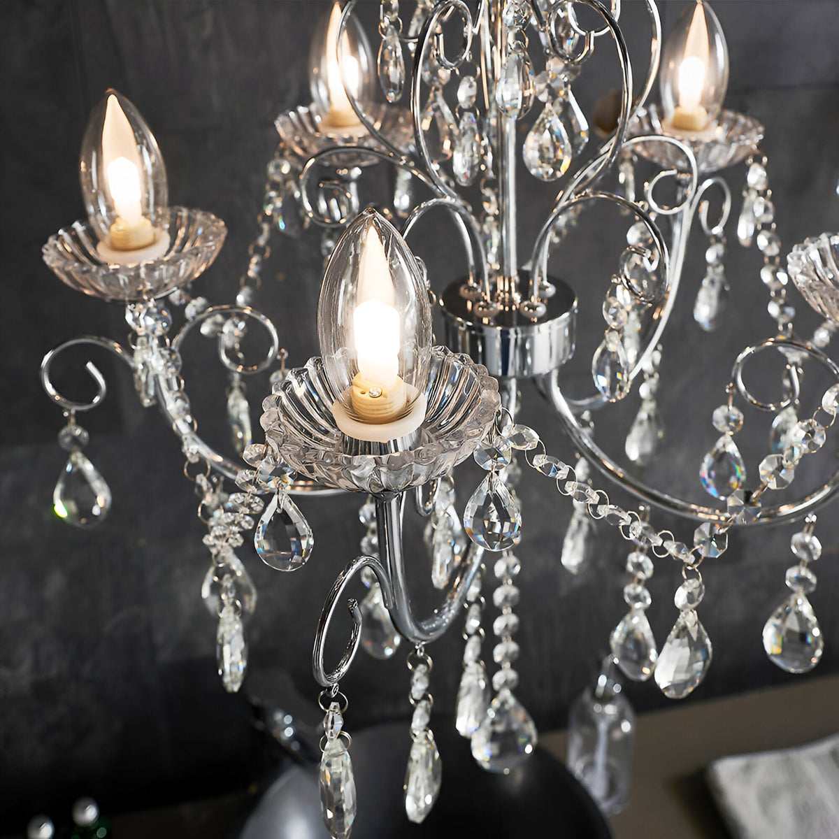 Endon Crandon 5 light bathroom chandelier in polished chrome with scrolled arms, clear faceted crystal droplets and lit candle bulbs against dark tiles.