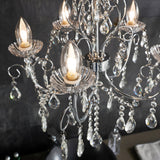 Endon Crandon 5 light bathroom chandelier in polished chrome with scrolled arms, clear faceted crystal droplets and lit candle bulbs against dark tiles.