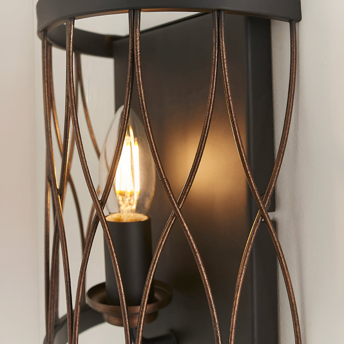 Close-up of the Endon Hulton wall light showing its crossed bronze wirework cage, matt black backplate and lit E14 candle bulb.